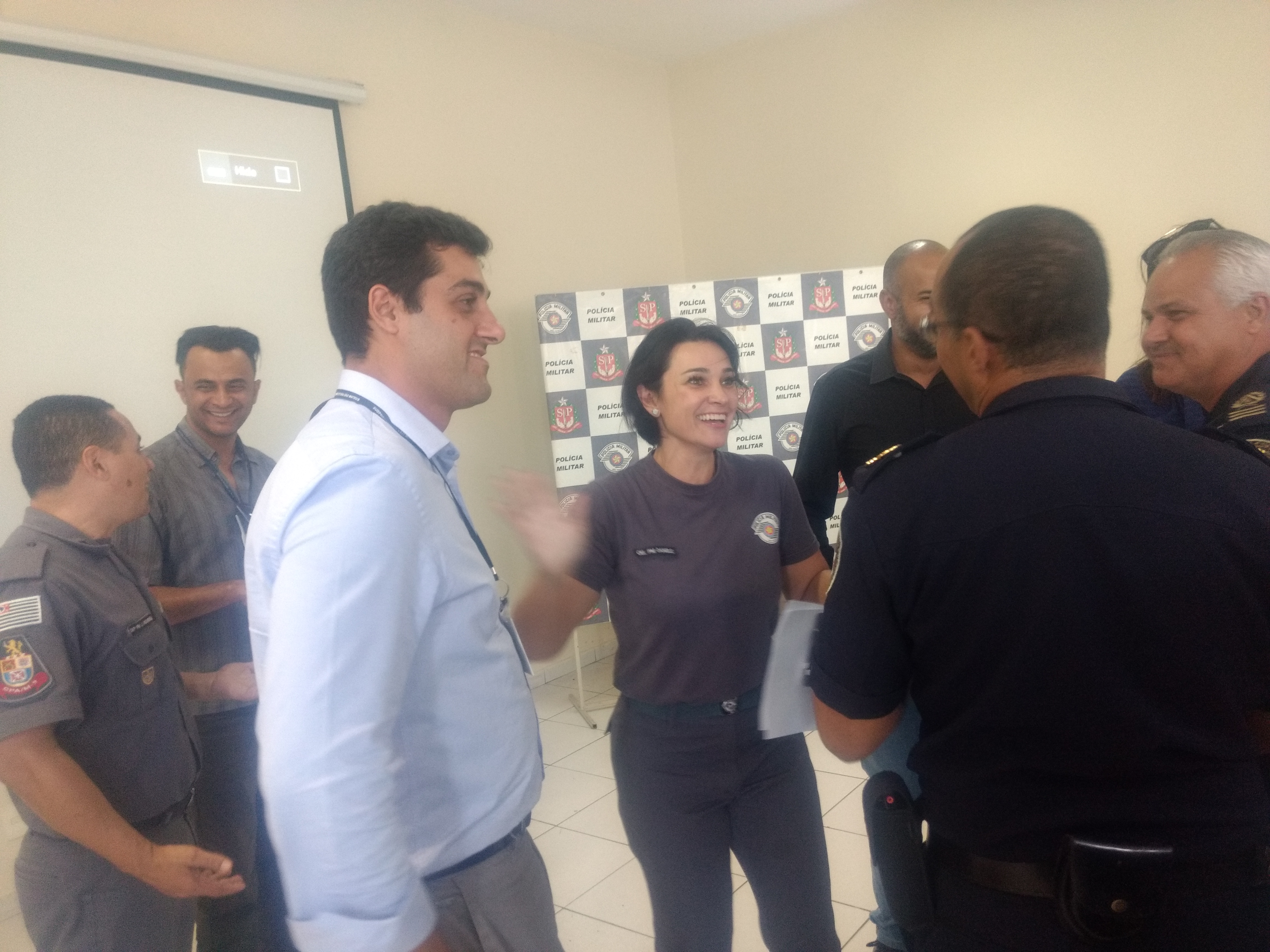 Ao centro, a comandante do CPA/M9, coronel Daniele, com a farda numa conversa informal e descontraída com um policial da Guarda Civil Metropolitana, que aparece de costas. Ela tem cabelos curtos e sorri, com a mão direita erguida, em movimento, pois esta parte está desfocada. O subprefeito de São Mateus observa, com leve sorriso. Ele usa camisa de manga comprida azul clara. À direita, além do GCM que dialoga com a coronel, há outro guarda civil, de cabelos grisalhos, que observa, com leve sorriso. Um pouco atrás, um participante da reunião, moreno, cabelo raspado e de barba, cujo rosto aparece cortado. Ao fundo, painel com a logomarca da Polícia Militar em vários quadros. Ao fundo, à esquerda, um PM fardado conversa com um homem que está com camisa listrada de preto e cinza, é moreno, está sorrindo e tem cabelo baixo..