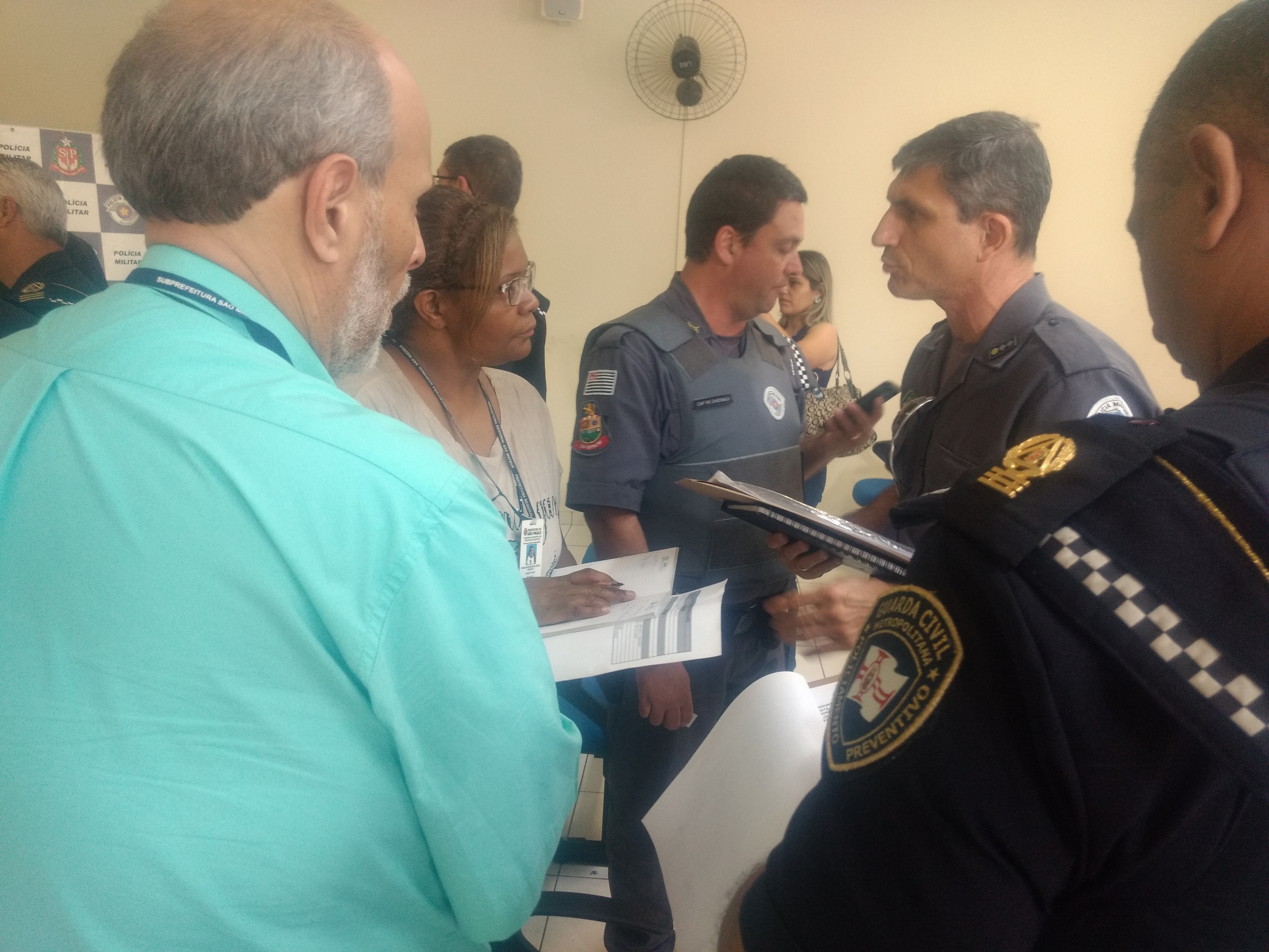 policial, com caderno em mãos, fala com a chefe de fiscalização da Subprefeitura de São Mateus, Ivani de Jesus, que faz anotações. Ao lado, o chefe de gabinete, De Angelis, presta atenção no que é dito. Mais atrás um policial com colete a prova de balas, olha no celular.