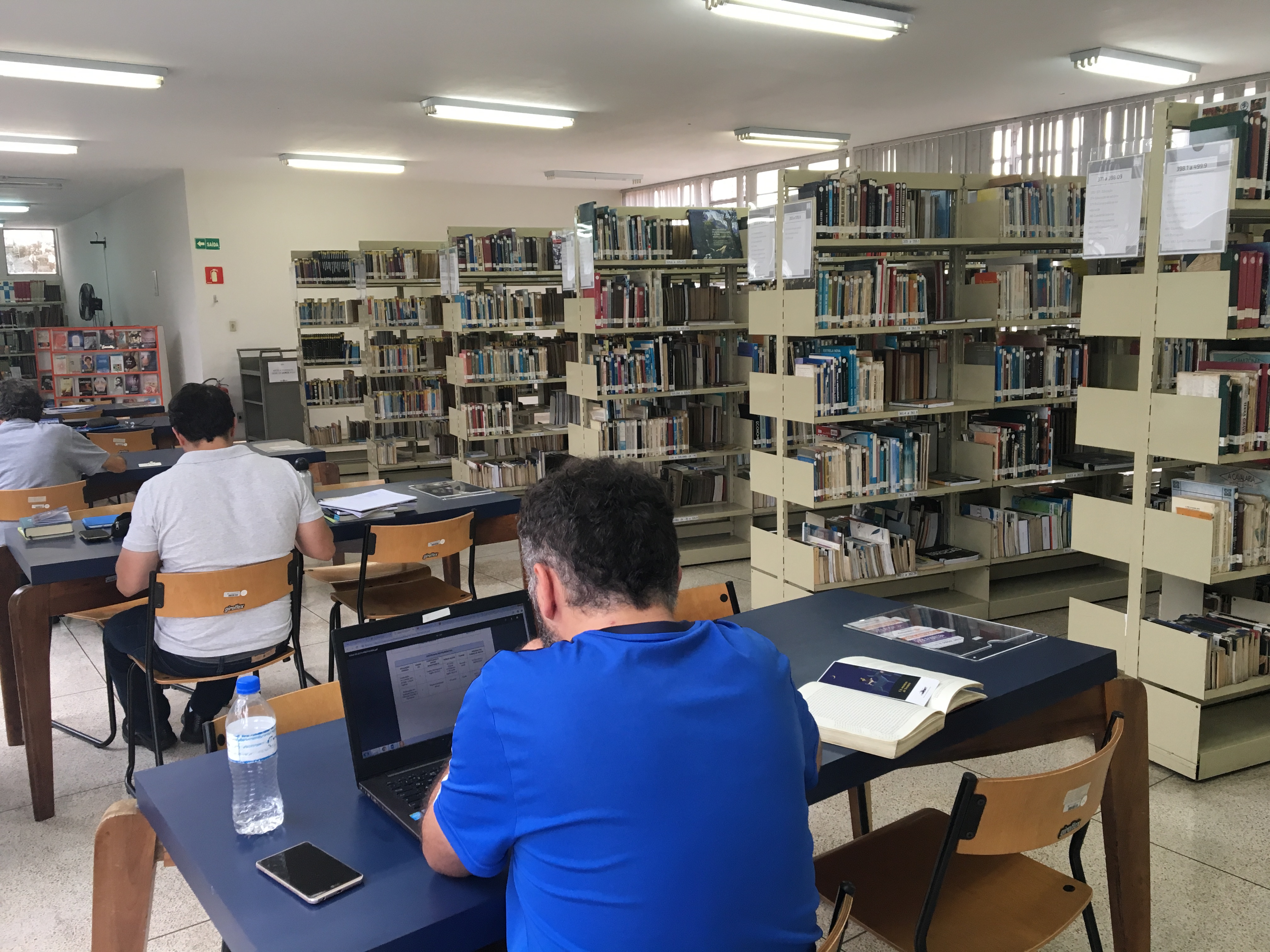 Três homens de costas, sentados em mesas diferentes da biblioteca. Dois estão lendo livros e um está lendo no notebook. Do lado direito tem seis estantes com livros