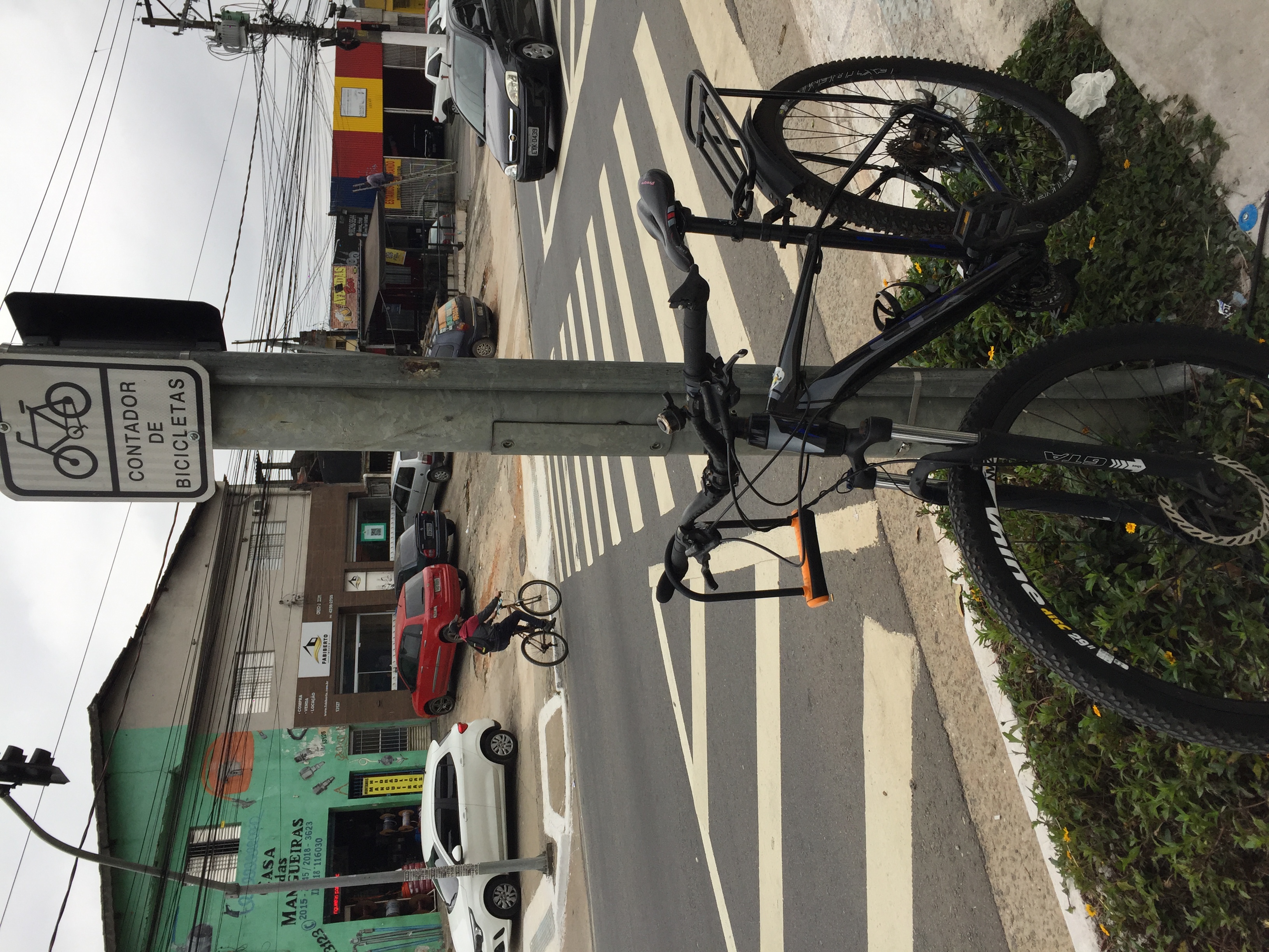 Bike está apoiada no poste metálico onde foi instalada a placa contador de bicicletas. Do outro lado da rua, um ciclista passa, fora da Ciclovia. 