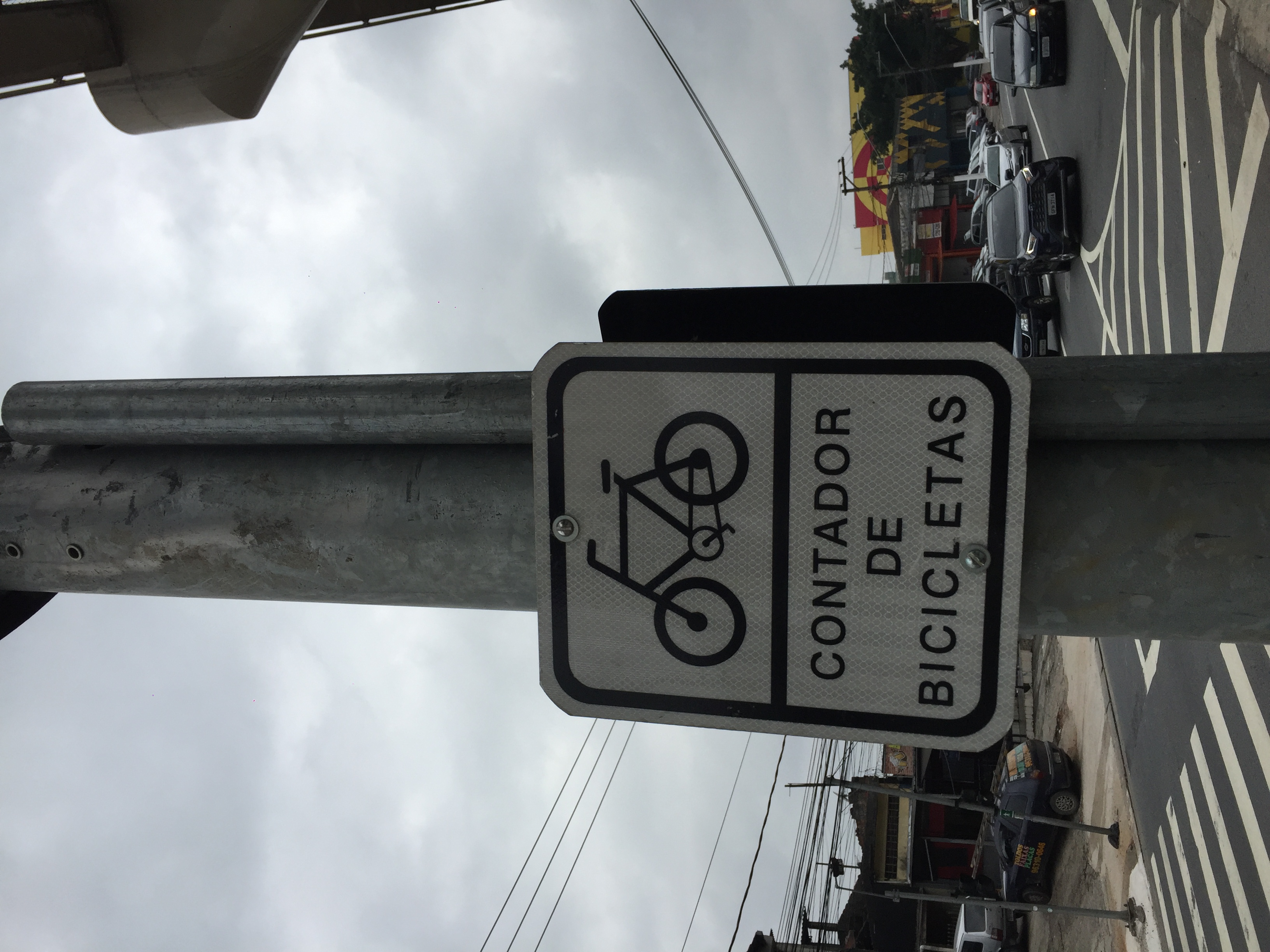 Placa oficial com fundo branco, letras  e desenho em preto indica onde há o contador de bicicletas. 