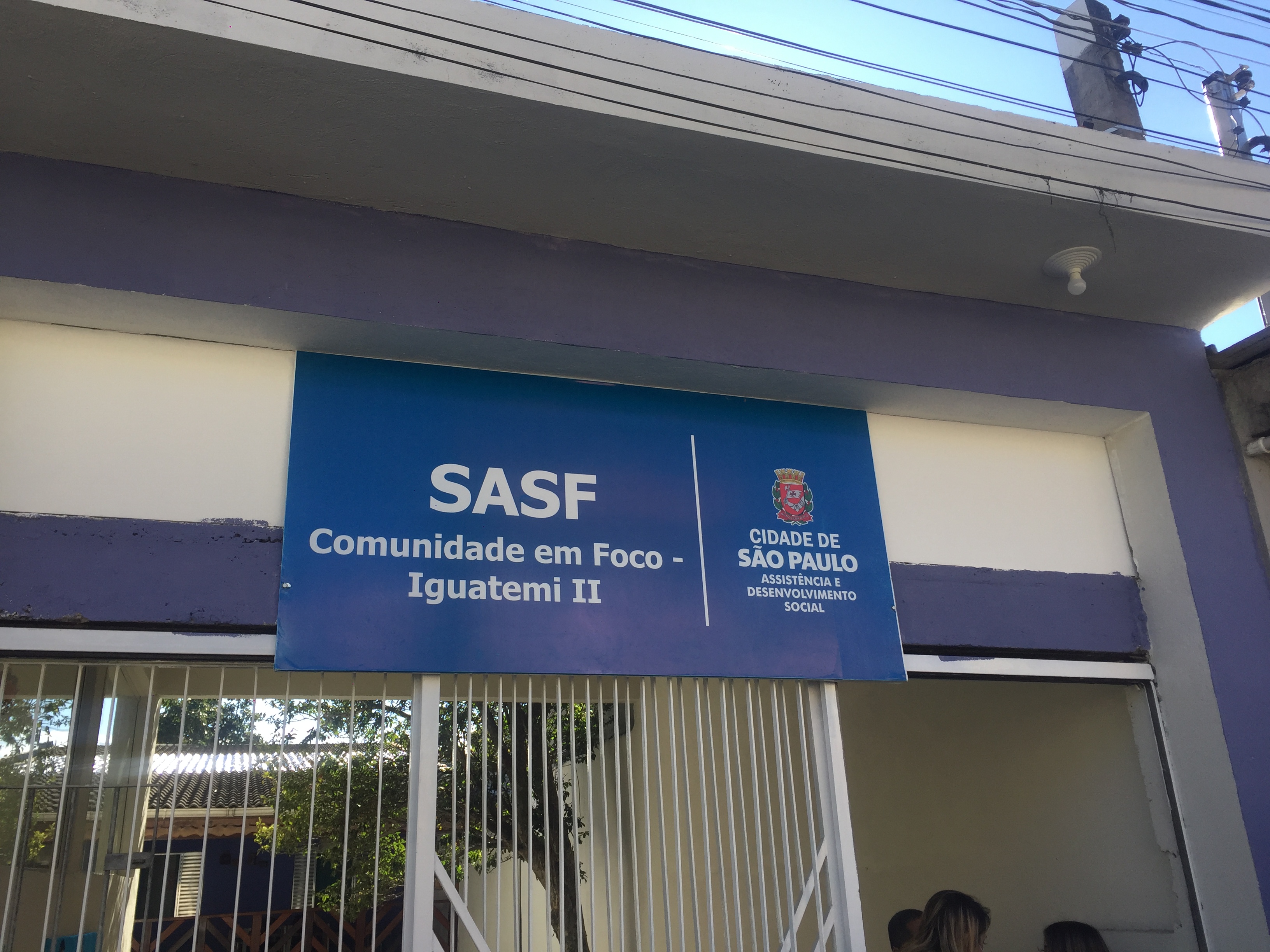 Placa com fundo azul e letras brancas, informa: SASF Comunidade em Foco Iguatemi II. À direita o logotipo Cidade de São Paulo, Assistência e Desenvolvimento Social
