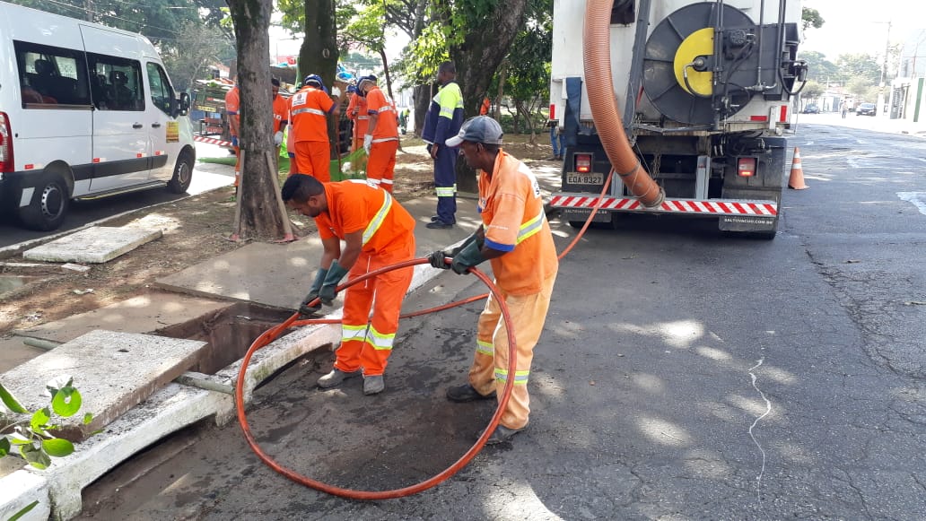 Imagem colorida, com equipe de limpeza mecanizada de bueiro, com duas pessoas usando uniforme laranja da Prefeitura, ao fundo outros trabalhadores executando outros serviços.