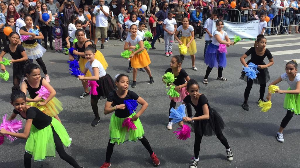 Na imagem mostram 17 crianças se apresentando com roupas coloridas no Desfile Cívico