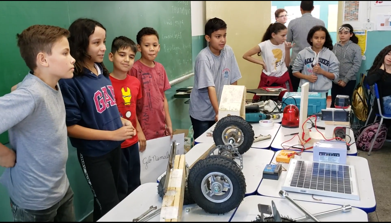 Na imagem 7 alunos estão perfilados na frente da mesa da sala de aula, expondo os materiais do carro. 