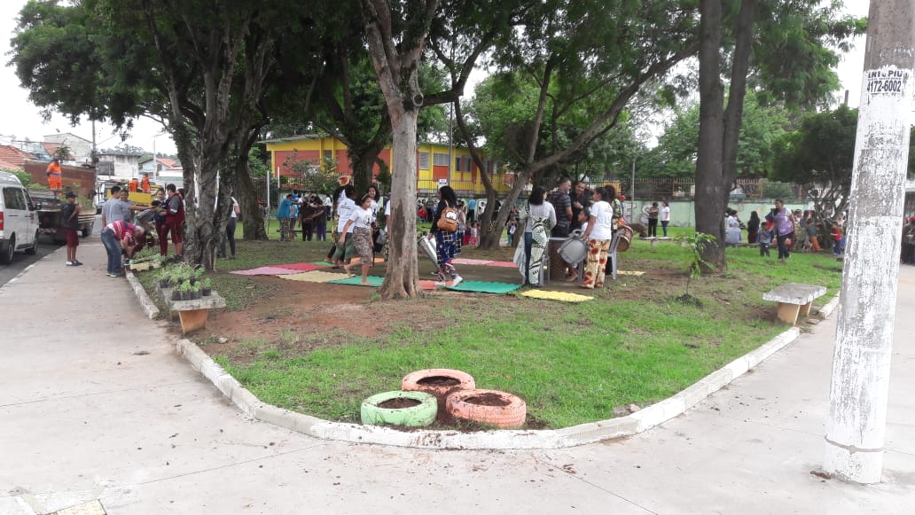 Na foto em plano aberto está a Praça  José Gualtierre Junior, cerca de 40 crianças e adolescentes com 10 professores e alguns funcionários de zeladoria, estão espalhados no parque. Algumas pessoas em pé e outros abaixados com mudas de planta na mão realizando o plantio no terreno do próprio parque ou em pneus velhos pintados 