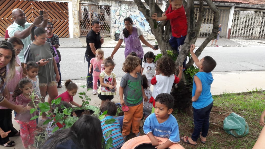 Na foto em plano fechado, cerca de 20 crianças estão com 5 professoras realizando o plantio de mudas de árvores, no canto direito da foto há uma árvore e uma criança sobe nela
