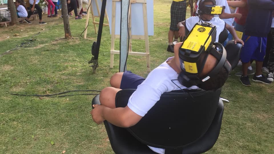 No plano principal da foto uma criança sentada em uma cadeira com rodinhas, a criança veste uniforme escolar e com os dois pés em cima da cadeira ele usa óculos de realidade virtual. Ao fundo da foto outra criança também de uniforme escolar, sentado em uma cadeira de rodinha, essa criança também usa óculos de realidade virtual.  