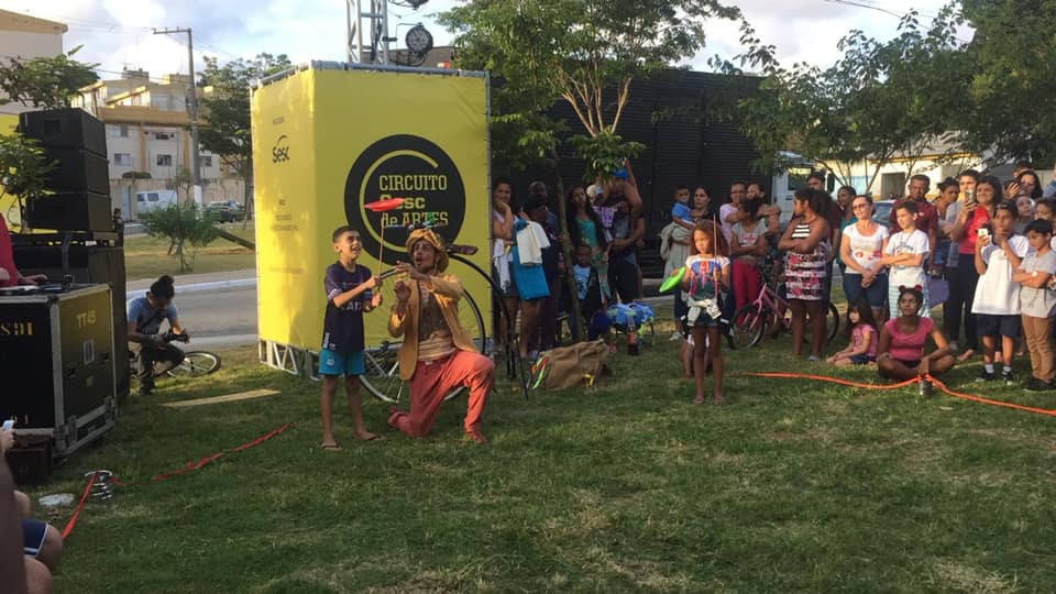 Em foto na Praça Almíscar cerca de 50 pessoas estão em roda crianças e adultos assistem enquanto um artista vestido de palhaço ensina uma criança a equilibrar um prato em palito. 