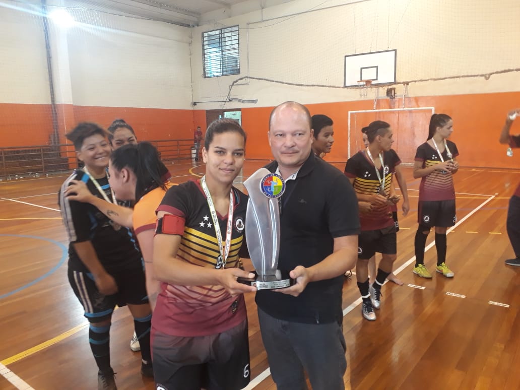 Na imagem 11 pessoas estão dentro da quadra, e uma mulher recebe um troféu do Jogos da Cidade de um homem em primeiro plano.