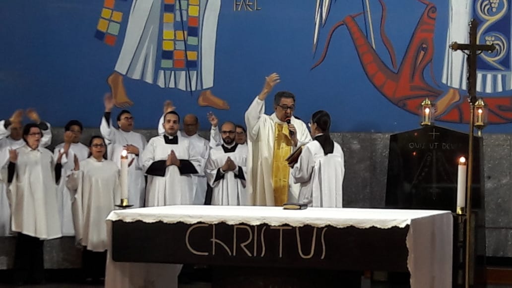 A imagem mostra o Padre Geraldo celebrando a missa juntamente com 11 pessoas ao fundo dentro da Catedral de São Miguel Arcanjo