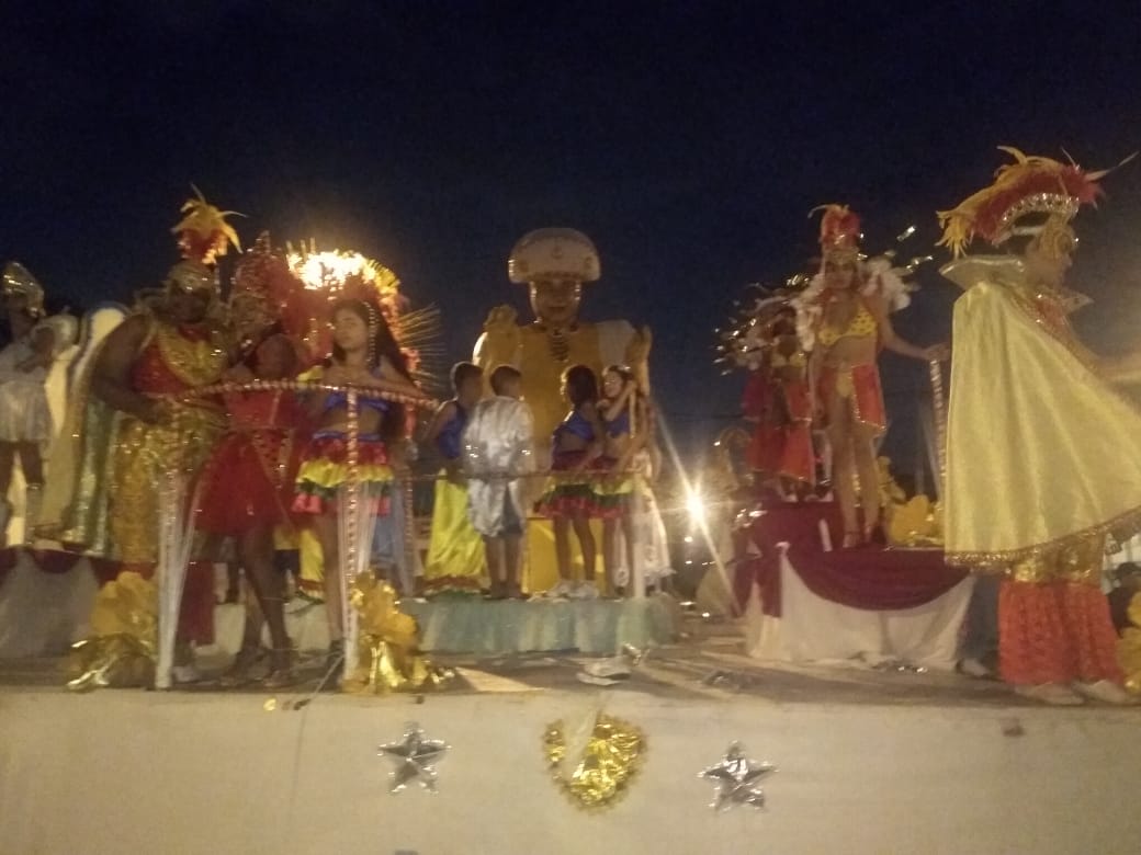 Na imagem um carro alegórico da escola de Samba Unidos de São Miguel, com cerca de 10 pessoas vestidas com alegorias de carnaval. A foto foi feita na Av. Dep. Dr. José A. Pinotti no período noturno.