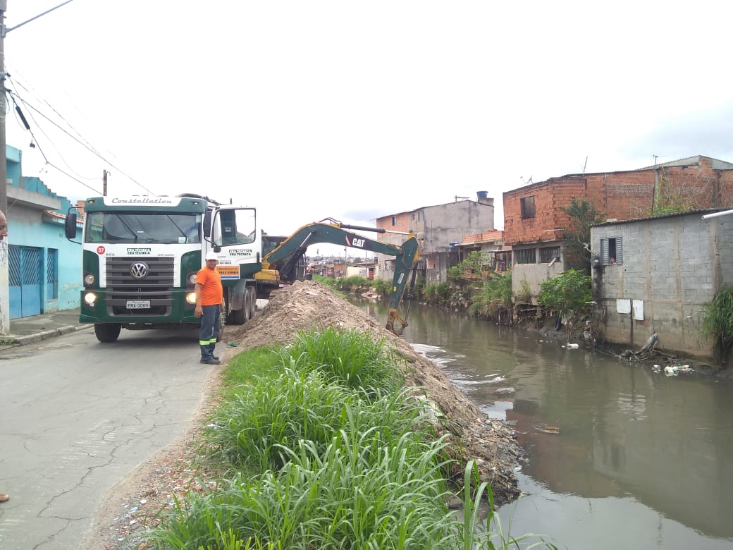 A foto mostra a limpeza no córrego na Rua Serra do Grão Mogol, um caminha está ao lado realizando a limpeza mecânica com um caminha. Um funcionário da Subprefeitura observa o serviço sendo realizado 