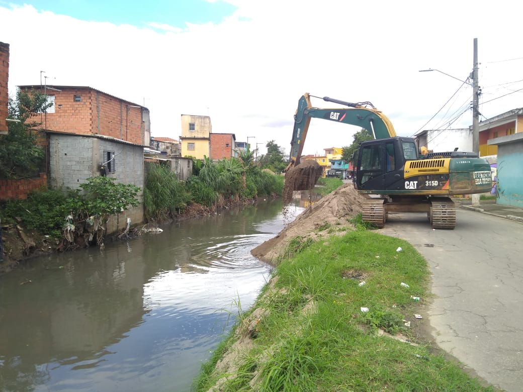 A foto mostra a limpeza no córrego na Rua Serra do Grão Mogol, um caminha está ao lado realizando a limpeza mecânica com um caminha