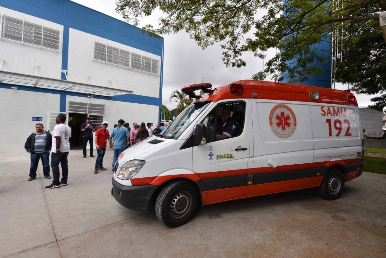 Em plano geral ao fundo a UBS Cidade Nova São Miguel, a esquerda alguns munícipes e na direita uma ambulância do SAMU.  