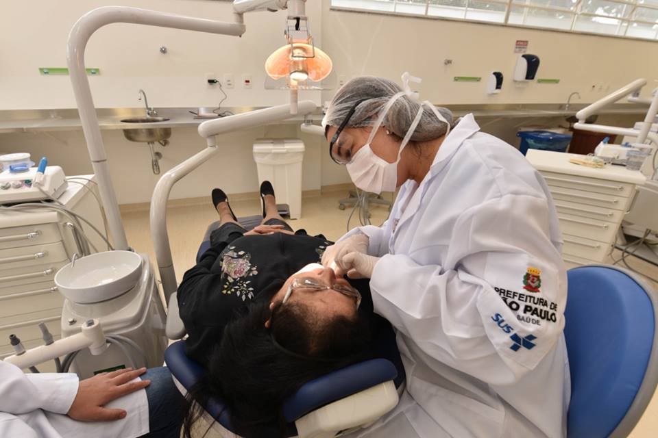 Na foto em um consultório odontológico, aparece a esquerda uma paciente deitada em uma maca, enquanto a dentista a direita atende a paciente.