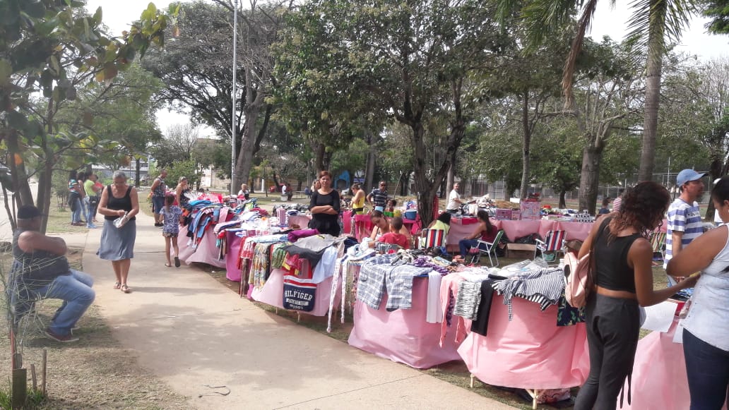 A foto mostra a praça craveiro do campo com diversas barradas cobertas com um tecido rosa bebe, existem roupas em cima de todas as mesas e pessoas estão próximas comprando-as 