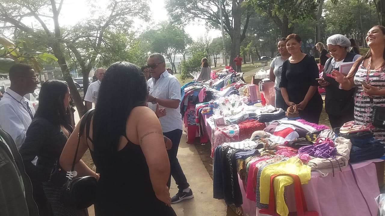 A foto mostra a praça craveiro do campo com diversas barradas cobertas com um tecido rosa bebe, existem roupas em cima de todas as mesas e pessoas estão próximas comprando-as 