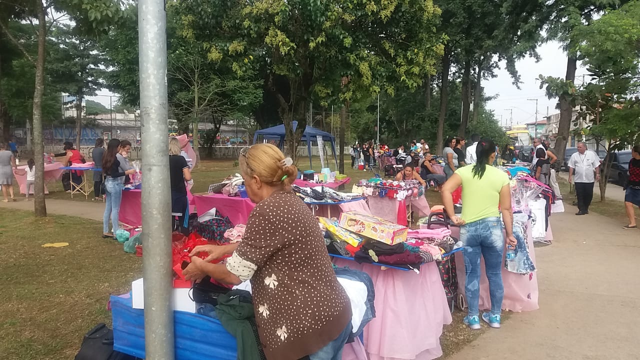 A foto mostra a praça craveiro do campo com diversas barradas cobertas com um tecido rosa bebe, existem roupas em cima de todas as mesas e pessoas estão próximas comprando-as 