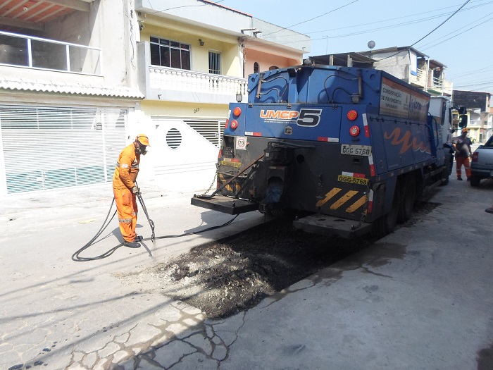 Trabalhador com uniforme laranja e equipamentos de proteção esguicha o material de reparos no asfaltos. O esguicho está ligado a um caminhão tanque, que está logo a seguir