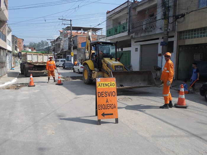 trabalhadores uniformizados com macacão laranja fazem o remendo no asfalto.  À direita, uma pá carregadeira, usada no serviço. A placa no chão avisa atenção desvio à esquerda 