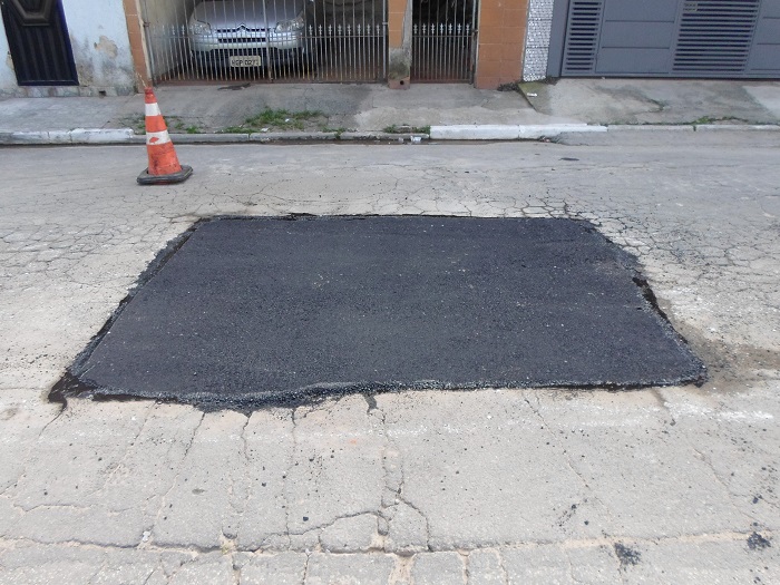 Uma mancha mais preta, retangular, no asfalto demarca o local em que o buraco foi tapado. À beira da guia, um cone laranja com duas listras brancas sinaliza a obra.