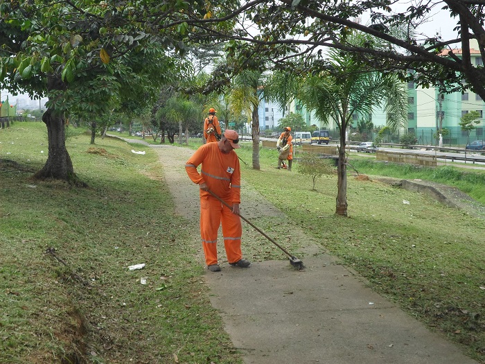 três trabalhadores fazem o corte de  grama. Um deles usa vassourão para limpar o passeio. 