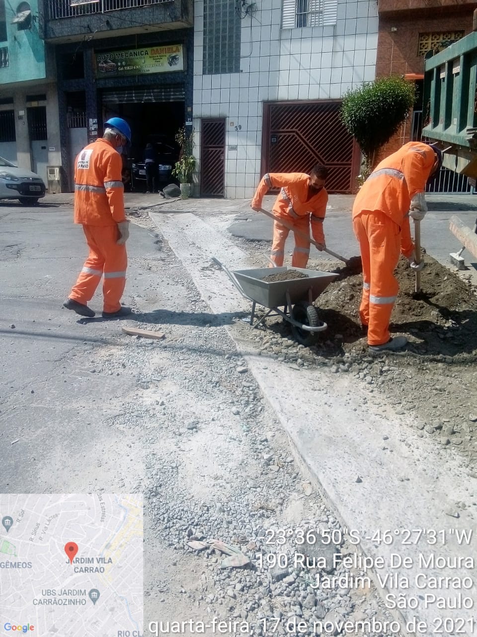 Três funcionários municipais, com uniforme laranja, preparam massa de concreto à beira de um sarjetão em fase final de reforma. 