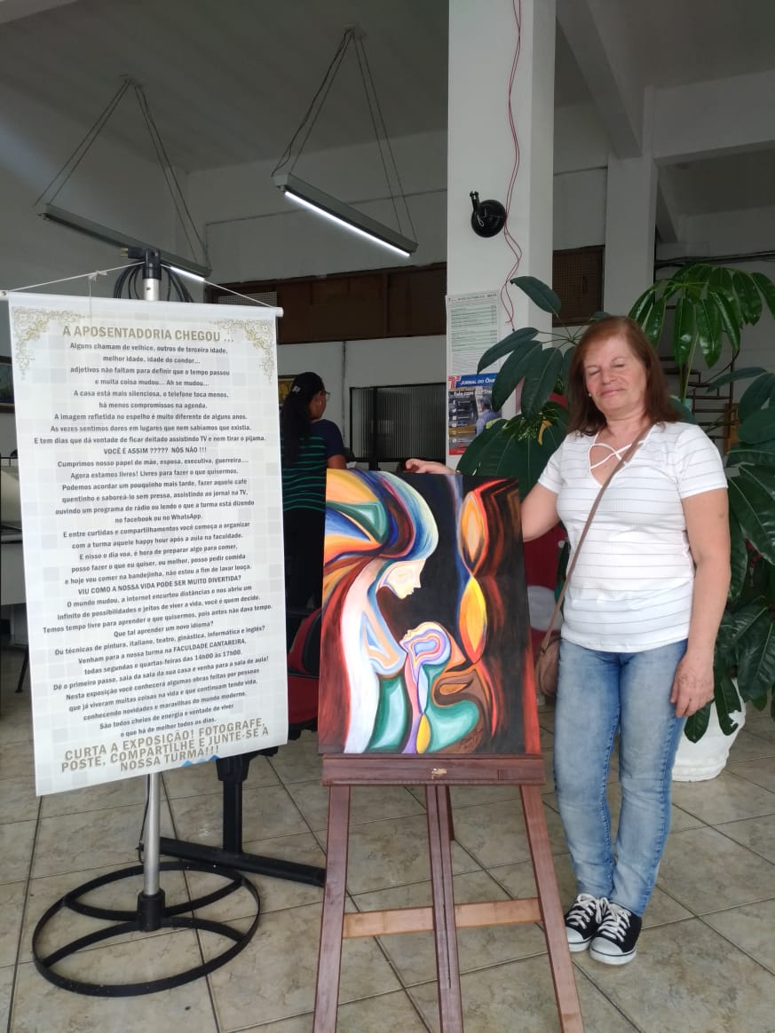 Imagem colorida, da artista plástica Cida Afonso, vestindo uma camiseta branca listrada, calça jeans, cabelos ruivos comprido, ao lado de um quadro pintado por ela a sua esquerda.