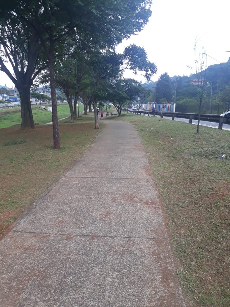 gramado limpo e podado, ao lado de uma avenida, protegida com alambrado metálico. Uma pista de caminhada ao centro