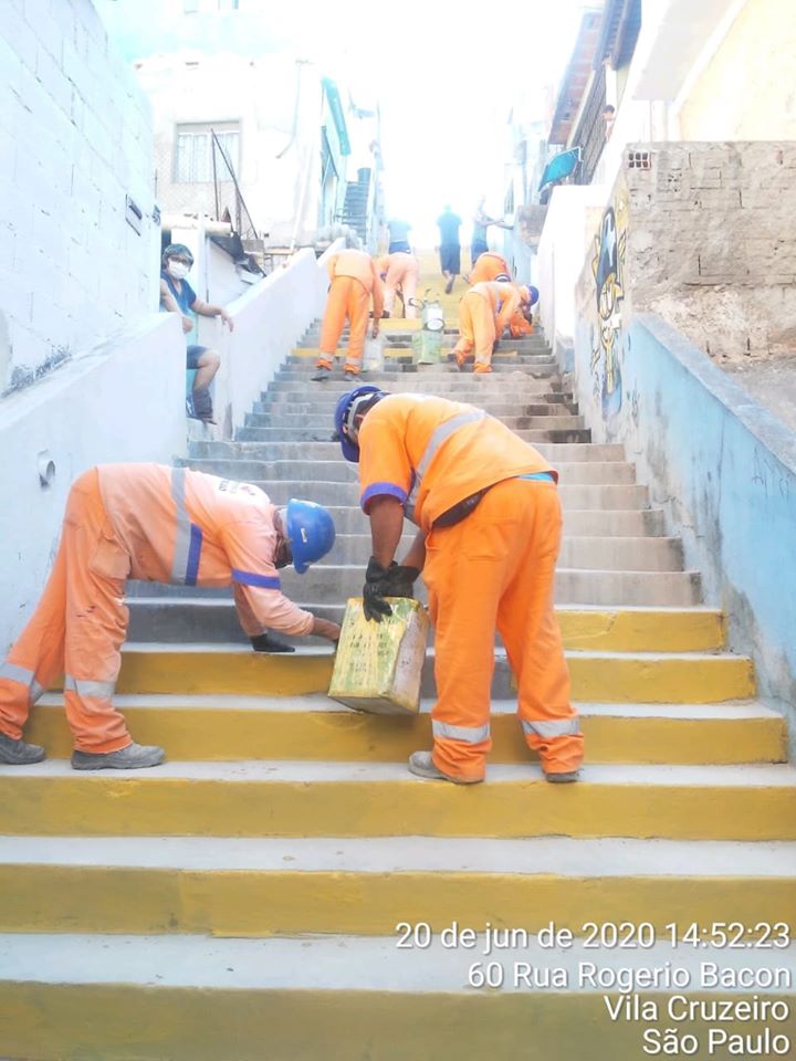 #PraTodosVerem visualiza-se trabalhadores, vestidos de uniformes da cor laranja, pintando uma escada de amarelo.