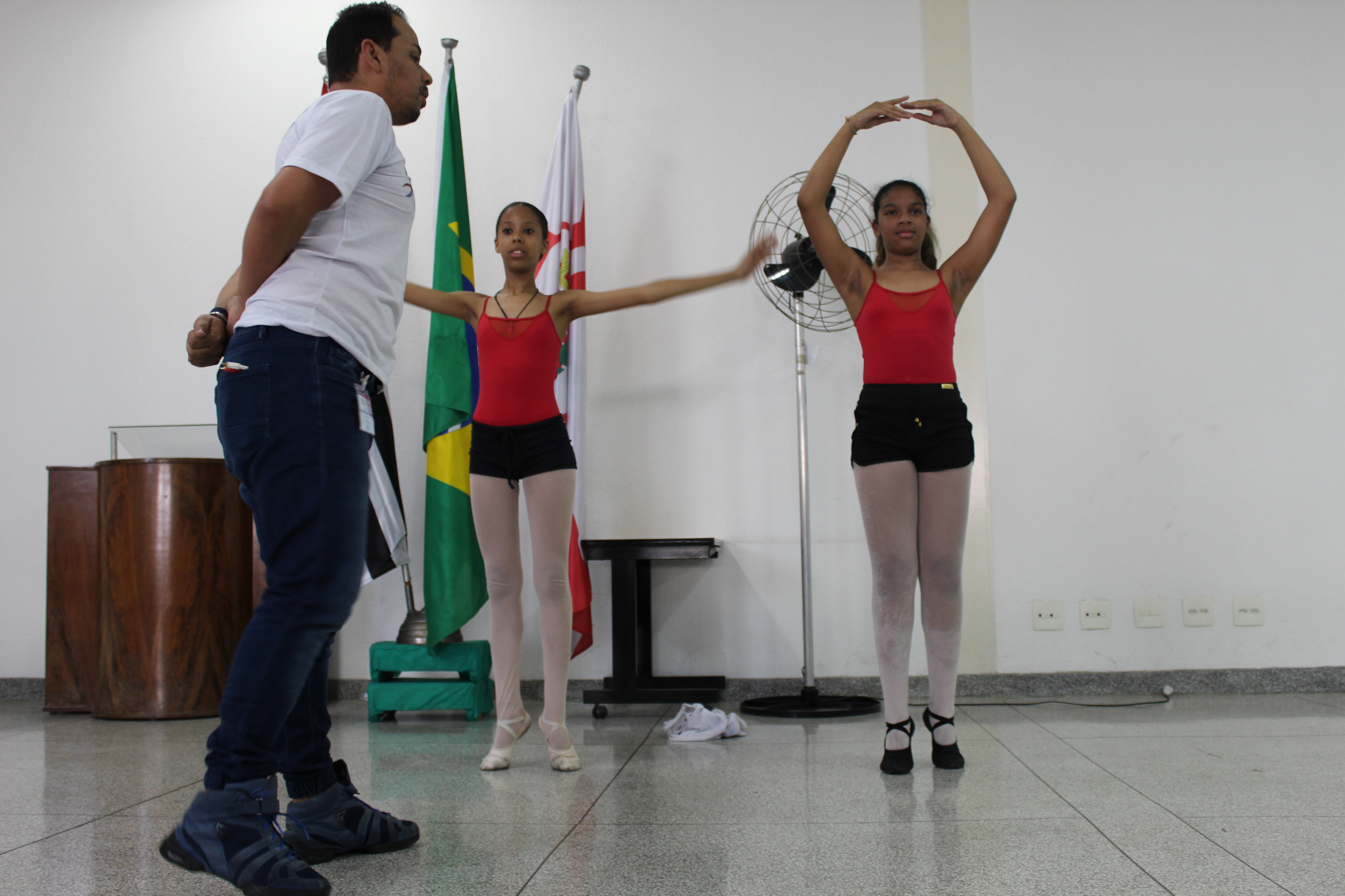 Duas meninas, com roupas de bailarinas, fazendo passos de Ballett enquanto o instrutor as orienta
