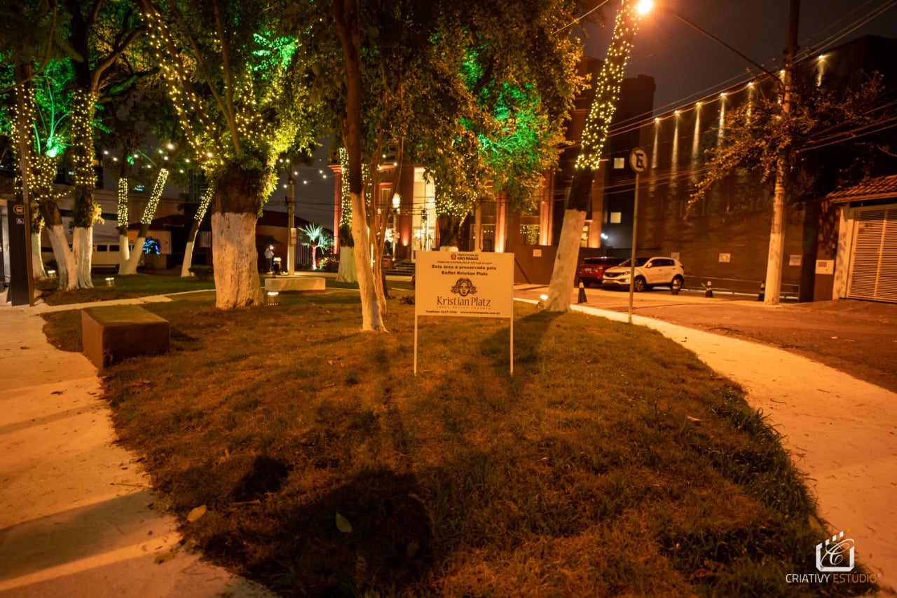 #PraTodosVerem visualiza-se a imagem de uma praça, decorada com luzes de led de cor amarela no tronco das árvores e verde nas folhas. Além disso, há uma placa com informações e logomarca do adotante.