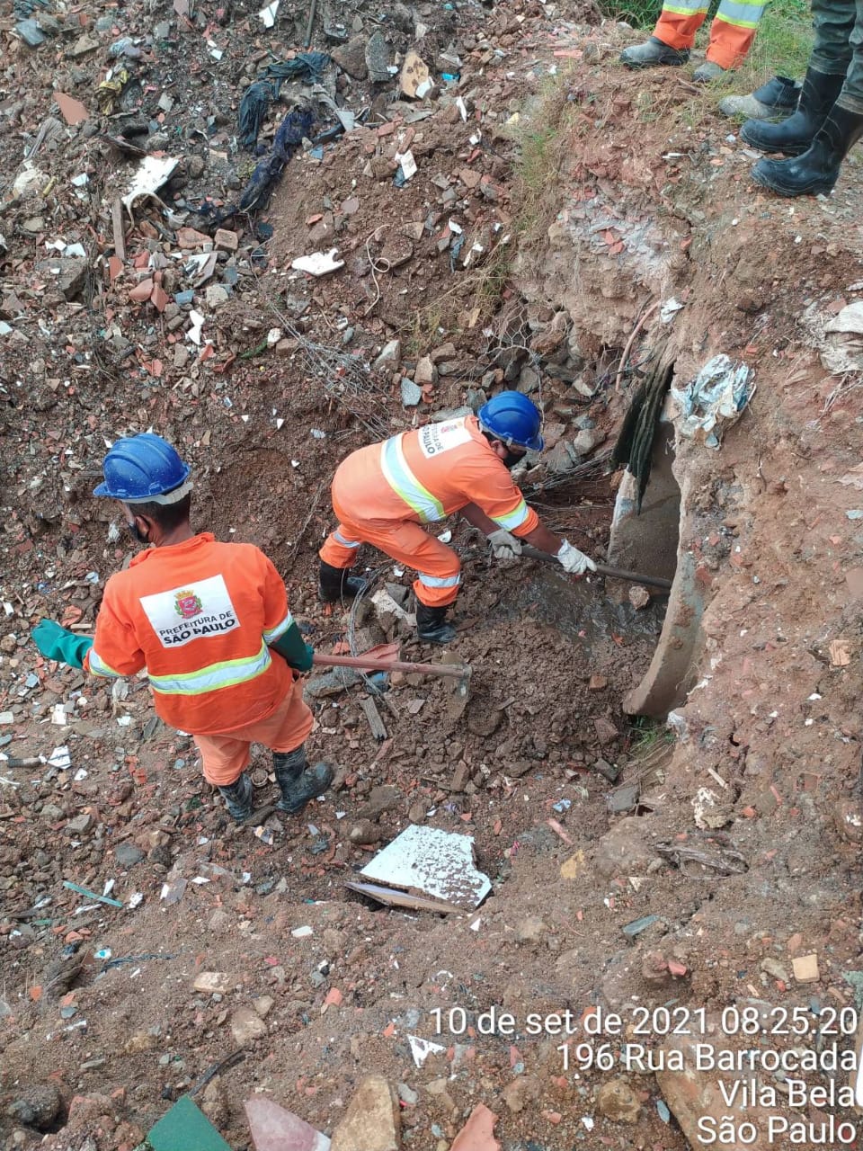 Dois homens, com uniforme laranja e capacetes azuis fazem a limpeza de um tubo de concreto, com entulho servindo de cobertura.  Rua Barrocada, Vila Bela. 