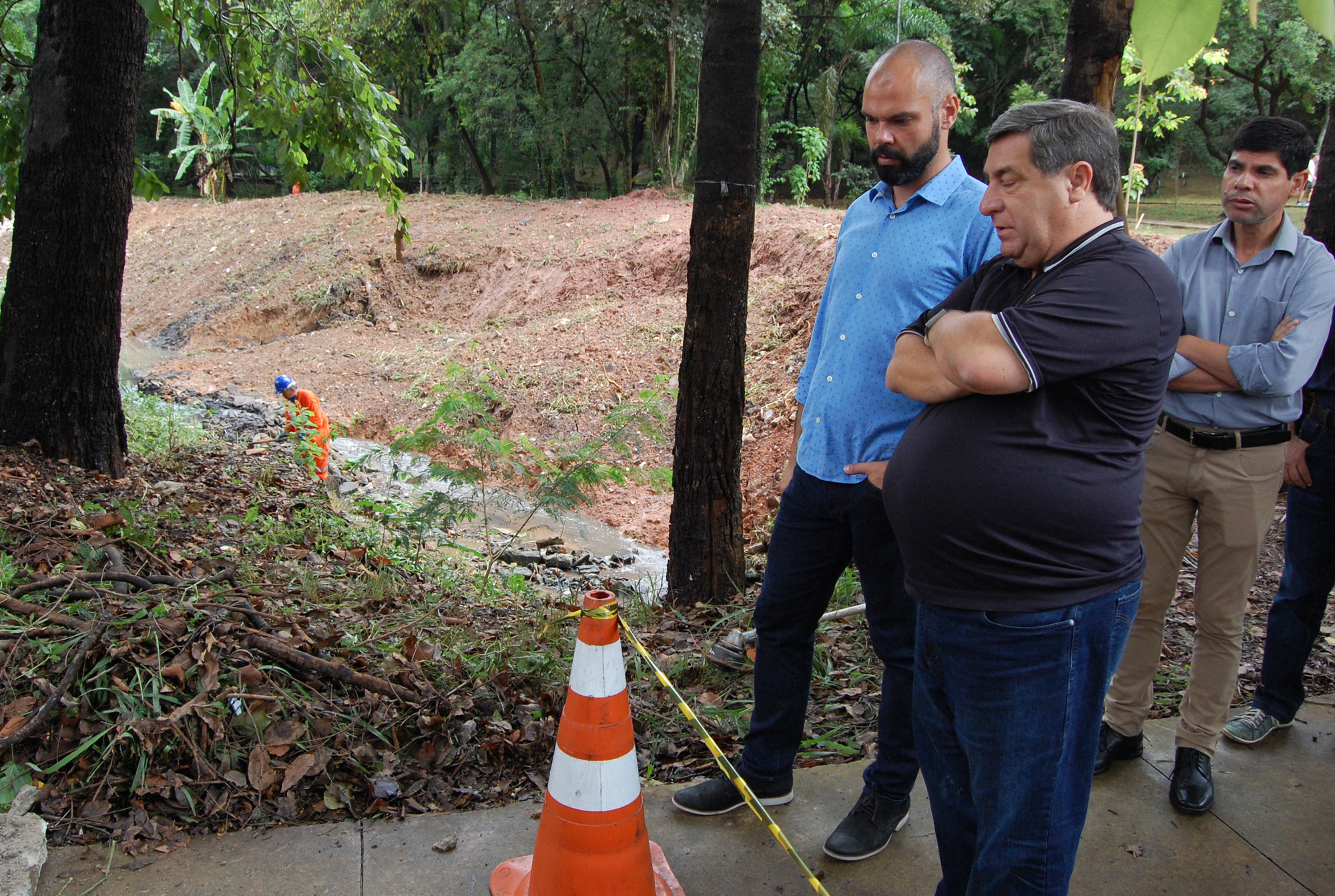 Foto do Prefeito Bruno Covas e do Subprefeito do Butantã, Paulo Vitor Sapienza, acompanhando a limpeza de um córrego. Temos ao fundo da foto um membro da equipe de limpeza na margem do córrego.