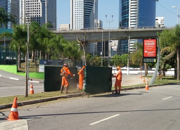 A imagem mostra uma avenida larga em primeiro plano. No centro da foto, funcionários de uniforme laranja cortam a grama de um canteiro - painéis verdes e cones são usados para isolar a área em que o trabalho acontece. Ao fundo observa-se uma ponte, árvores e prédios.