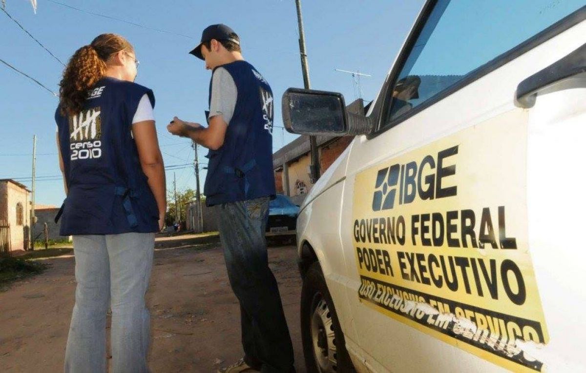 A imagem mostra dois funcionários do IBGE realizando a coleta de dados para o Censo. No lado direito da imagem há um carro branco, com um adesivo amarelo de identificação do IBGE. Já no lado esquerdo, uma mulher e um homem  observam o aparelho usado pelos Agentes Censitários. Ambos são funcionários do IBGE e vestem um colete azul que os identifica como tais. O cenário constitui-se de uma rua de terra, com casas e postes presentes em ambos os lados. 