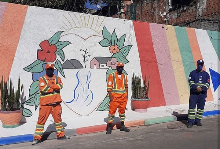 A imagem mostra três funcionários em pé em frente a calçada. Atrás tem um muro com uma arte colorida - um coração decorado com flores, um sol, uma paisagem e listras coloridas. Na calçada, também, foram colocados dois vasos de planta.