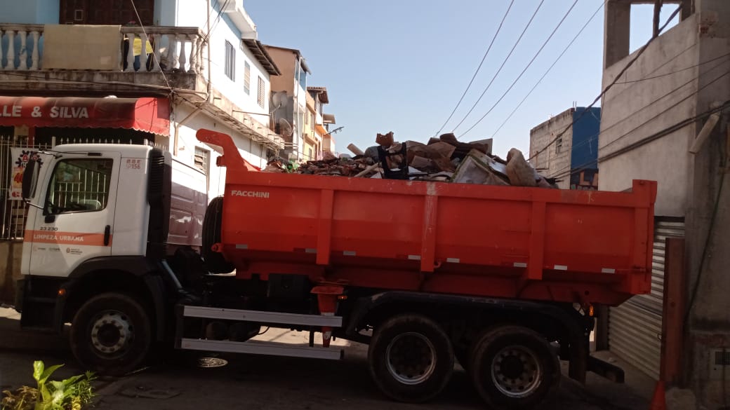 Foto de caminhão de Cata-Bagulho percorrendo ruas da comunidade da São Remo. O caminhão está com bastante material em seu compartimento. A rua possui uma grande quantidade de casas, que estão localizadas ao fundo da imagem.