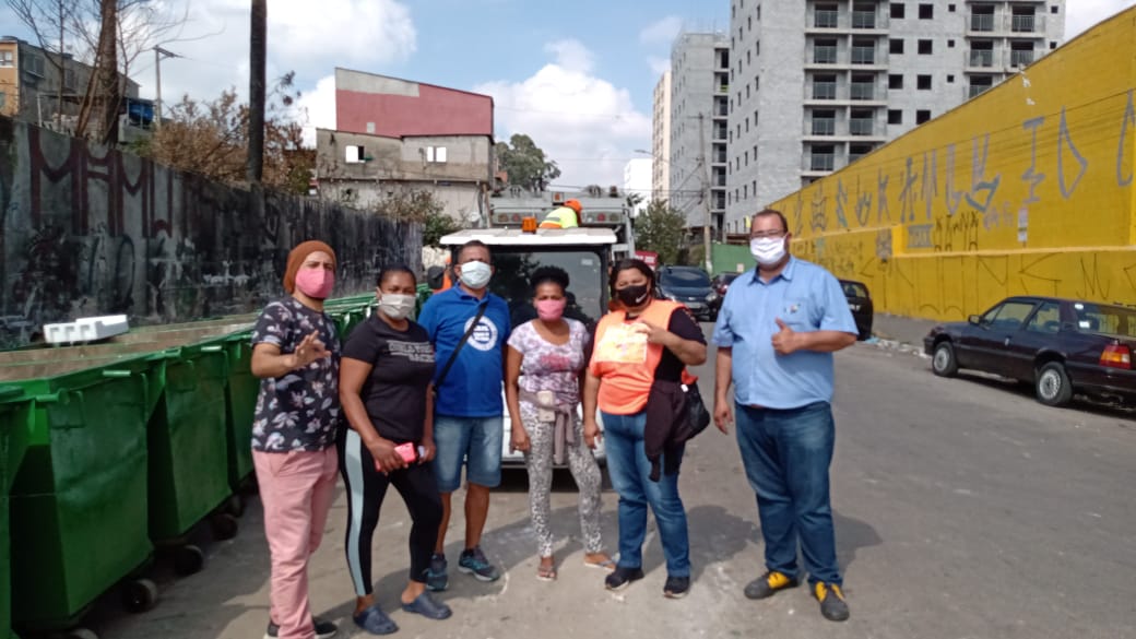 No centro da imagem estão os líderes comunitários da São Remo. Ao lado esquerdo lixeiras estão posicionadas. Ao fundo dos líderes um veículo de recolhimento de lixo pode ser parcialmente visto. 