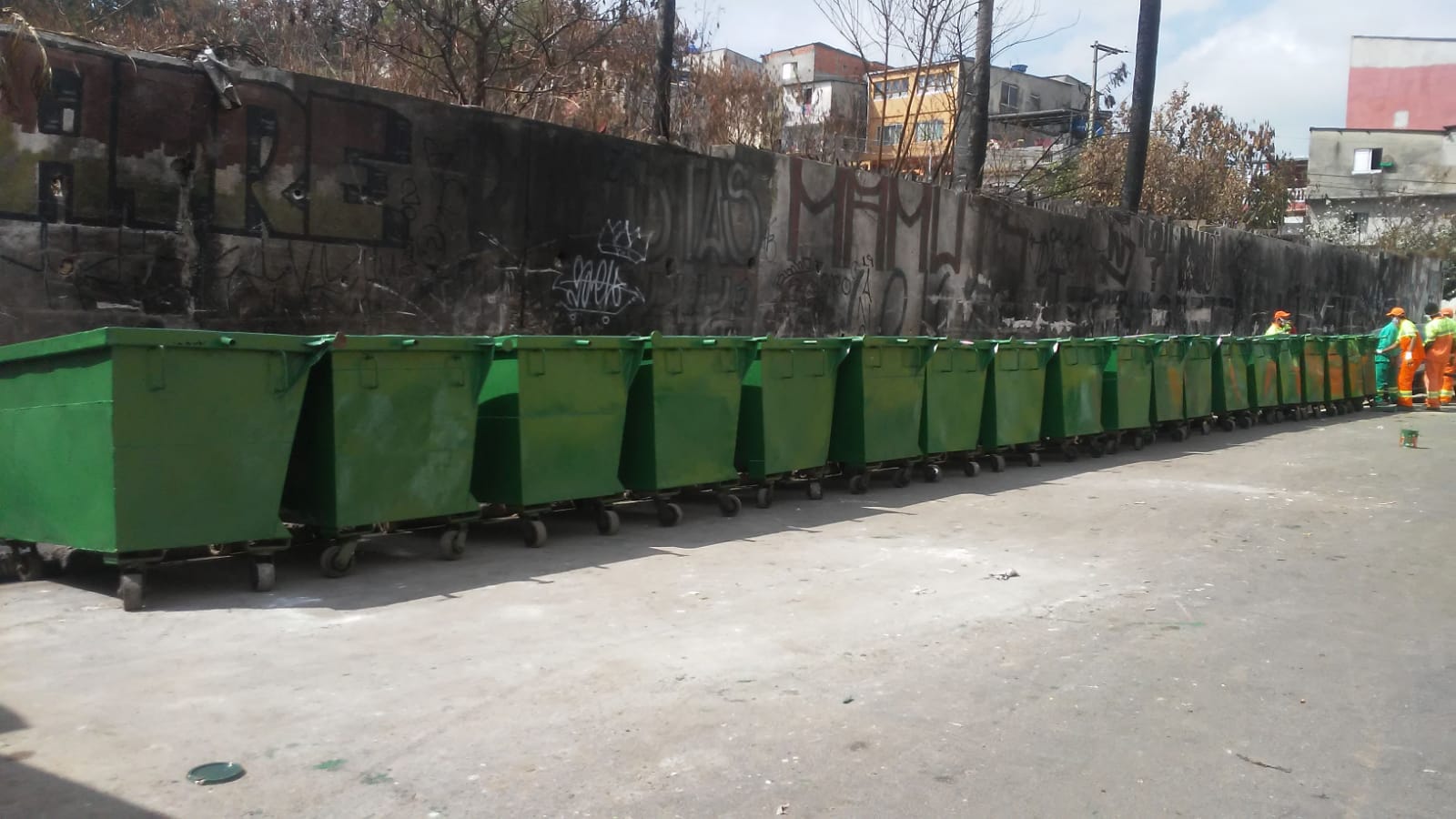 Foto de várias lixeiras, de grande porte e cor verde, sendo instaladas em rua da comunidade São Remo. Ao fundo funcionários uniformizados da Prefeitura de São Paulo podem ser avistados. 