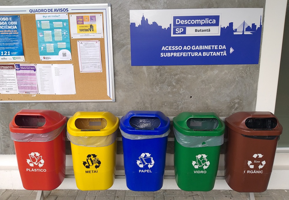 Foto de cinco lixeiras de reciclagem, que estão próximas à entrada da Subprefeitura, todas as lixeiras possuem o símbolo da reciclagem e, abaixo do símbolo há uma indicação por escrito do material que pode ser descartado, sendo cada lixeira própria ao descarte de um material. Cada uma das lixeiras possui uma cor própria para o descarte de um material específico, a vermelha é para os plásticos, amarela para os metais, azul para os papeis, verde para vidro e, marrom para materiais orgânico.