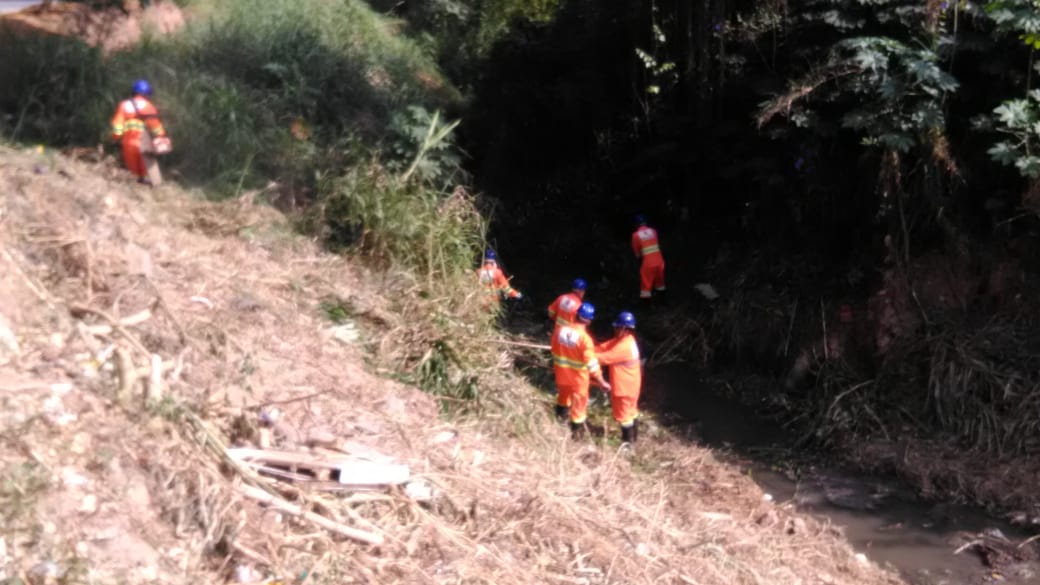 	Foto da equipe de limpeza manual de córrego adentrando ao Córrego da Passagem Grande. A grama no entorno do córrego está cortada a equipe composta de seis homens, todos vestidos de macacões laranjas, entram no córrego para fazer a limpeza. É perceptível que os homens seguram ferramentas para realizarem o trabalho.