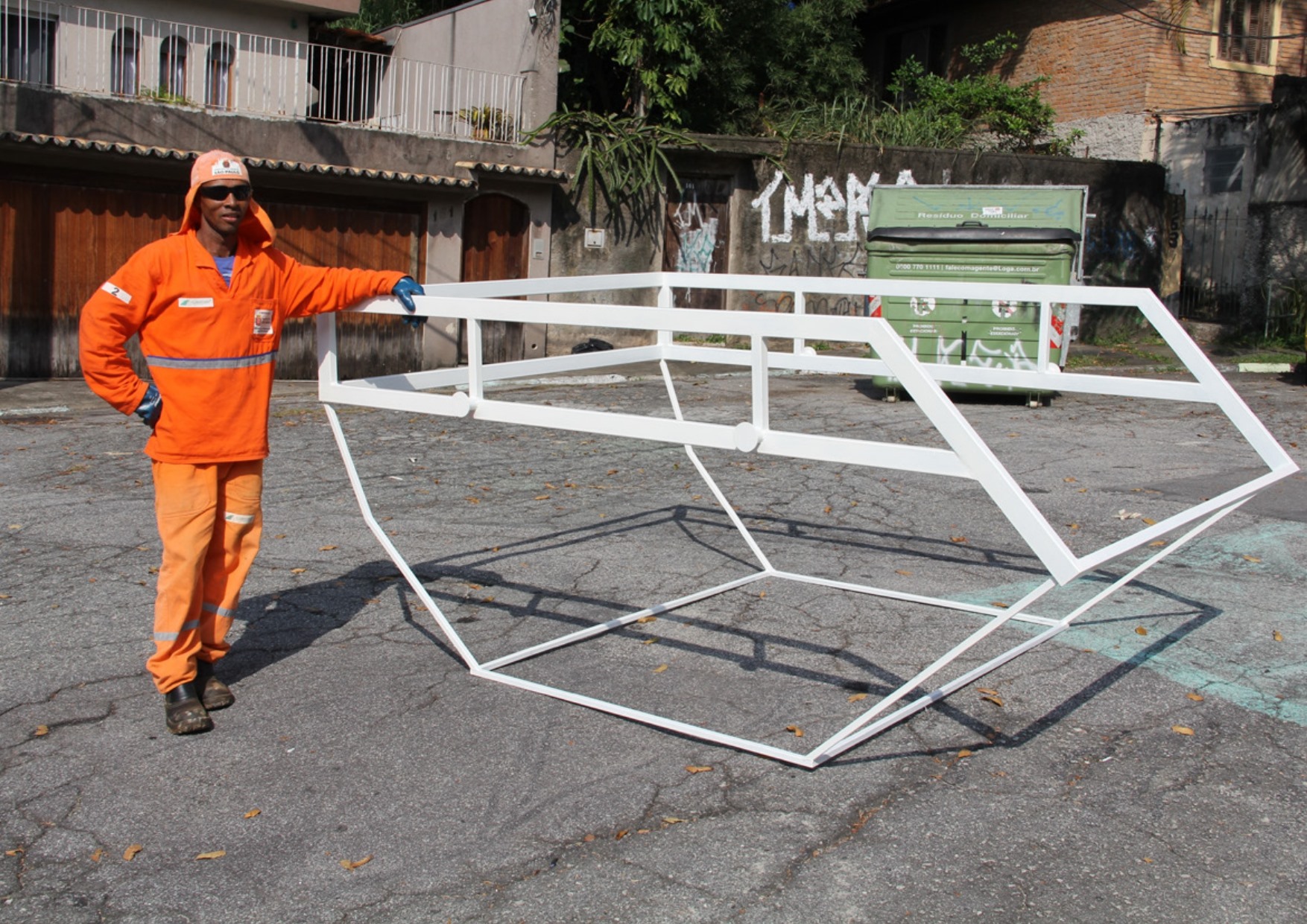 Representação de uma caçamba com espaços vazados e linhas de metal branco no meio da rua. Funcionário da Subprefeitura vestido de laranja posando ao lado da escultura apoiando uma das mãos.