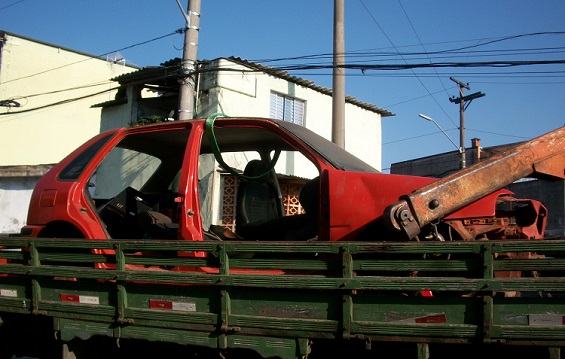 Foto de uma carcaça de carro, modelo Gol de cor vermelha, sendo recolhido encima de um caminhão.