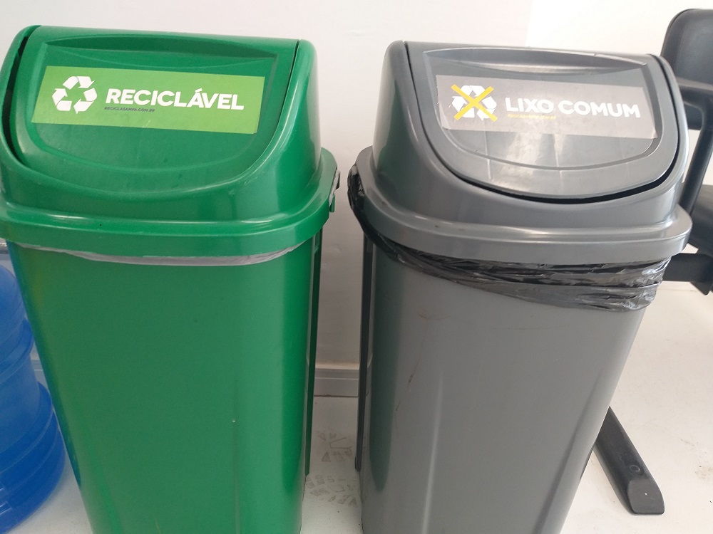 Foto das lixeiras do Recicla Sampa implantadas nas salas da Subprefeitura, uma da cor verde para lixos Recicláveis e outra da cor cinza para lixos comuns. Ambas as lixeiras possuem adesivos indicando o material próprio para o descarte em cada uma das lixeiras.