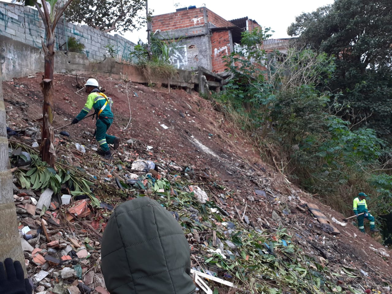 Foto de funcionários da Subprefeitura do Butantã trabalhando na remoção do lixo no talude. Um funcionário está sendo segurado por uma corda. Vemos outro funcionário que, está com a mão encostada em uma árvore enquanto olha para o funcionário preso com a corda. No chão há diversos tipos de lixos,  porém há uma predominância de restos de tijolos. No canto inferior direito da imagem temos outro funcionário da prefeitura trabalhando na remoção.