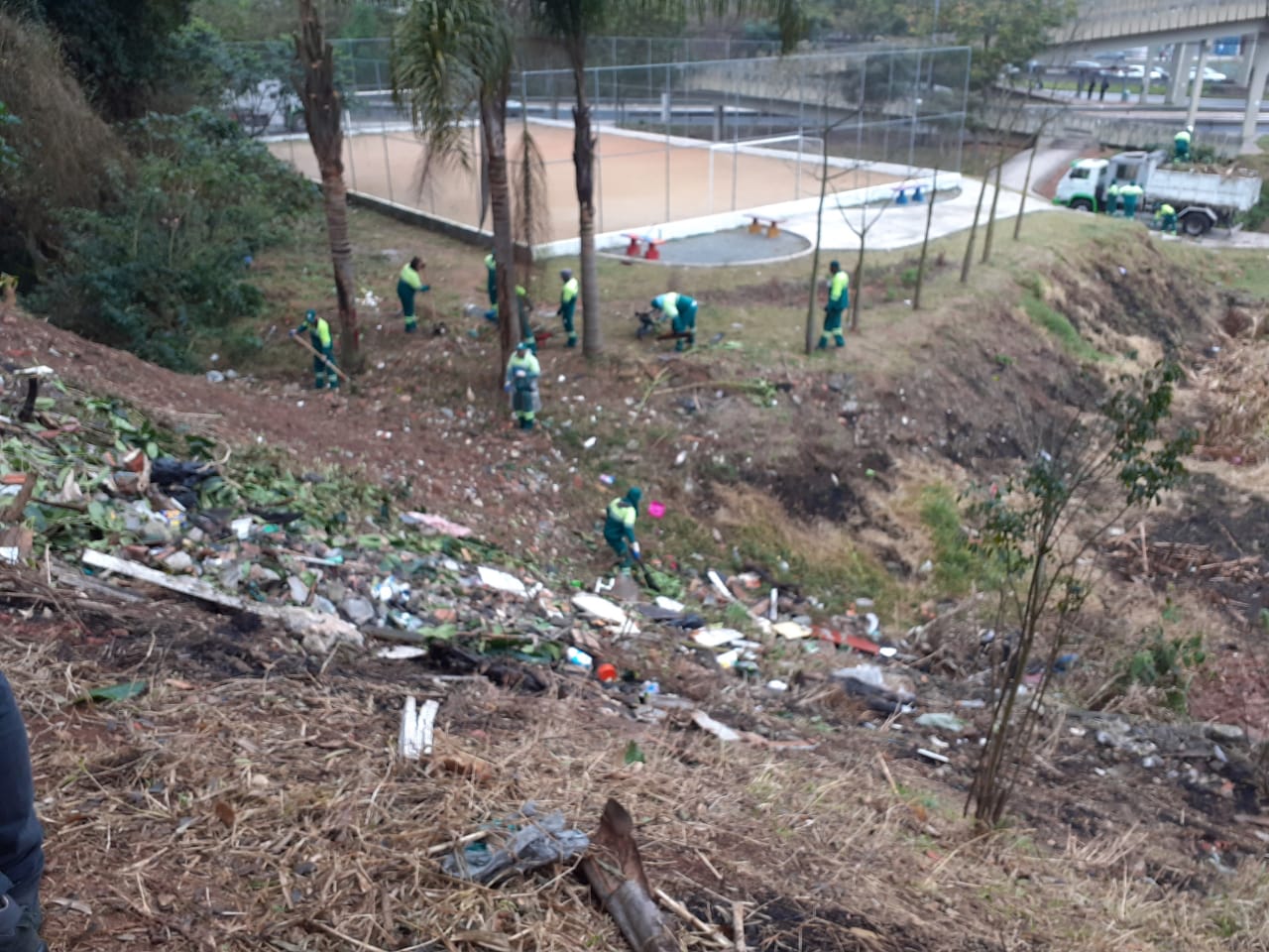 Foto dos funcionários da Subprefeitura trabalhando na parte inferior do talude, na parte superior existe uma grande quantidade de lixo. Ao fundo da imagem o caminhão da equipe está parado em uma rua.