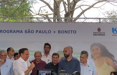 Foto do ato de entrega da revitalização da Praça Santos Coimbra, pelo programa SP+Bonito. O governador João Doria fala ao microfone, enquanto demais autoridades observam. Na foto foi capturada parte de uma filmadora profissional, o que indica a presença da imprensa no evento. Estão presentes também o Prefeito Bruno Covas, o Secretário Edsom Caram, o Subprefeito do Butantã, Paulo Vitor Sapienza e o Subprefeito do Jabaquara, Arnaldo Faria de Sá.
