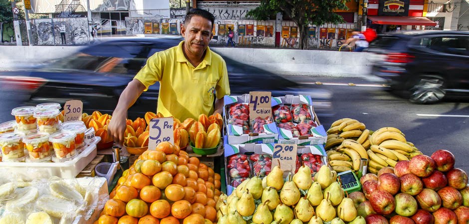 Foto de um comerciante e sua barraca de frutas, que está instalada numa calçada, próxima a uma avenida. Na barraca temos diversas frutas como pêra, maçã, banana, caqui, morango, laranja, melão e potes de saladas de mix de frutas. Encima das frutas plaquinhas de papelão indicam os preços, uma máquina de cartão de crédito pode ser vista. Ao fundo carros e bicicletas transitam pela avenida.